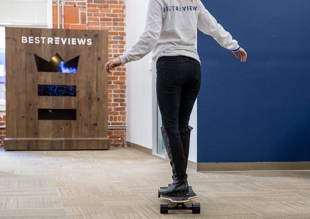 A person wearing black pants rides a longboard across a large, open office space.