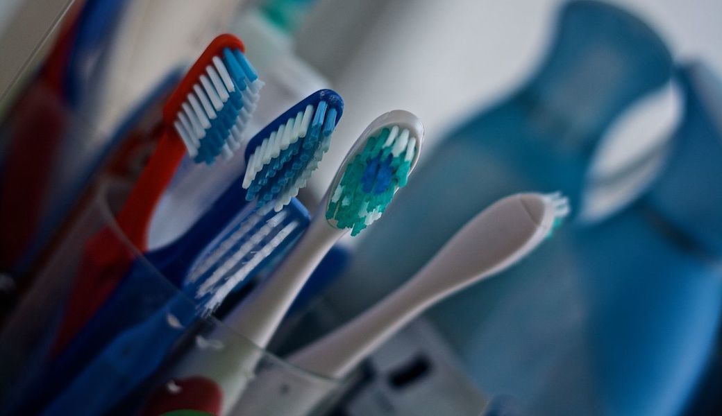 A collection of multi-colored toothbrushes in a glass container with blurry cleaning products in the background